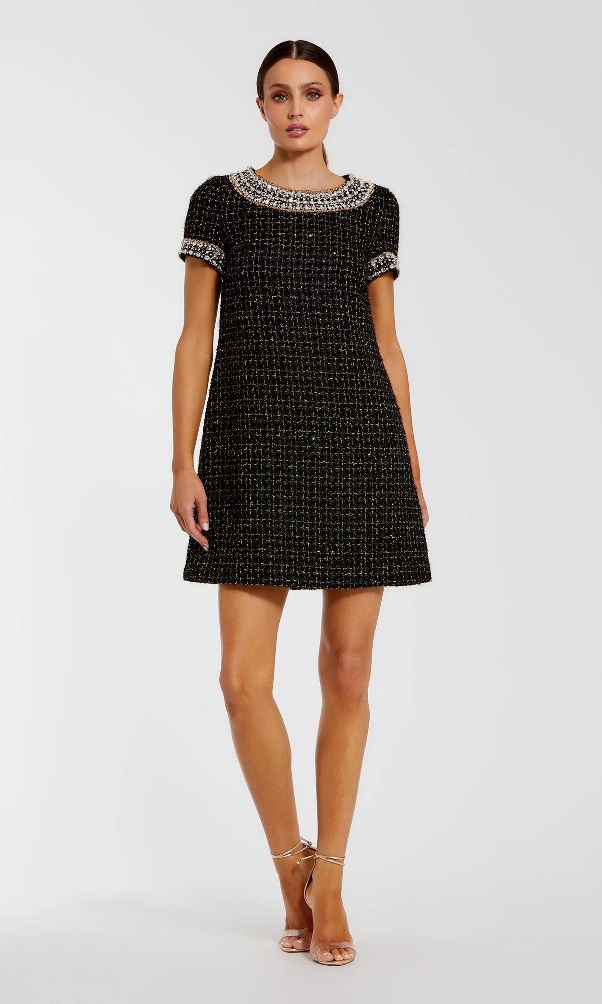 A woman stands against a plain white background wearing the Short Semi-Formal Dress: Mac Duggal 37075, a black gold mini dress with short sleeves and an embellished collar and cuffs adorned with rhinestone beading, paired with strappy heels.